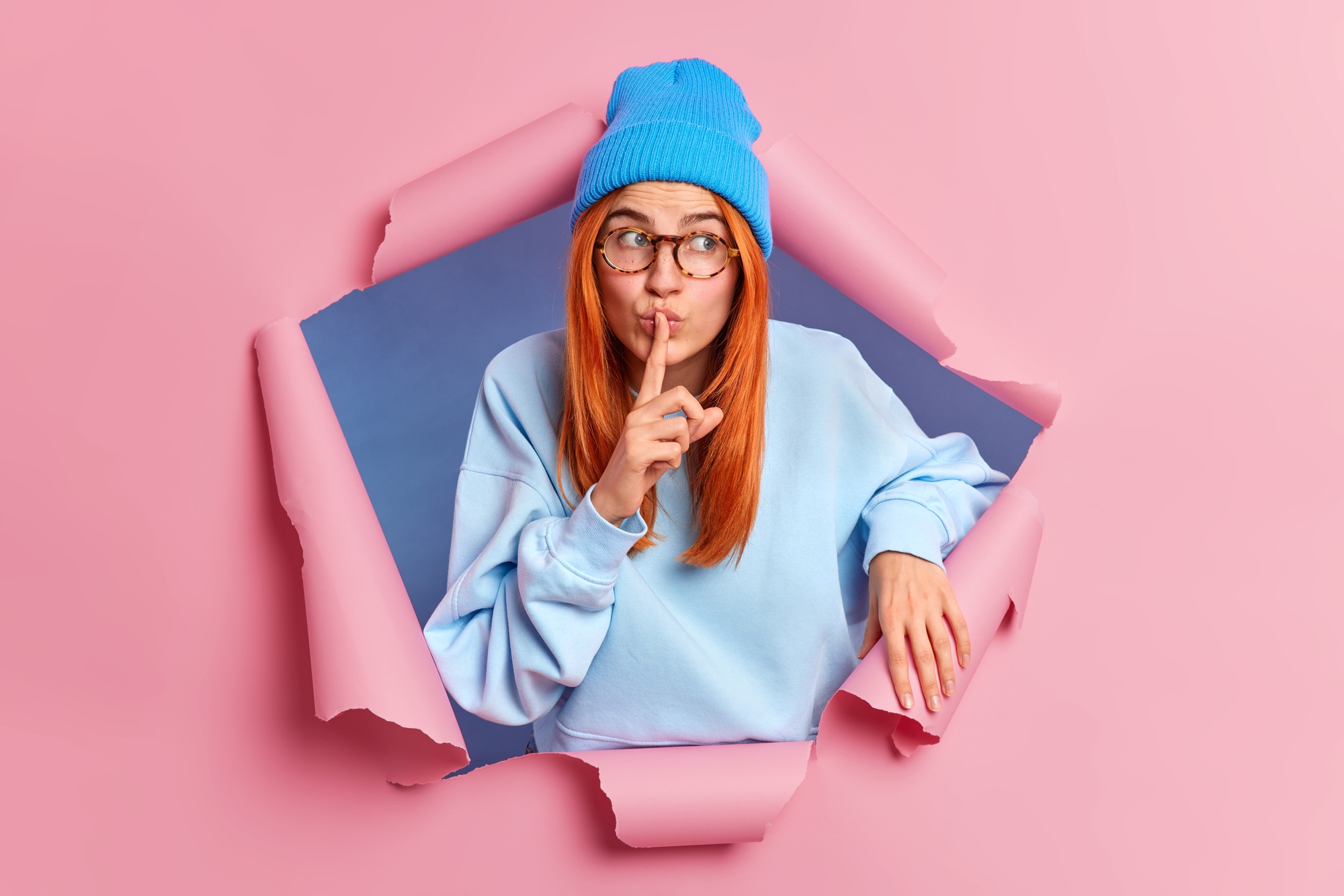 Mysterious redhead woman makes silence gesture asks not to tell her secret wears blue hat and sweate